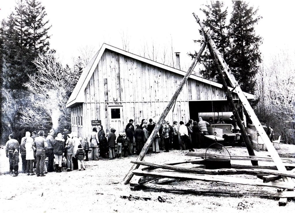 A large crowd of people form a line in front of a wooden building with a slanted roof. They line up for a pancakes and fresh maple syrup made at the demonstration. In front of the crowd, a pot is held over a fire using a rope attached to a tree logs propped up.