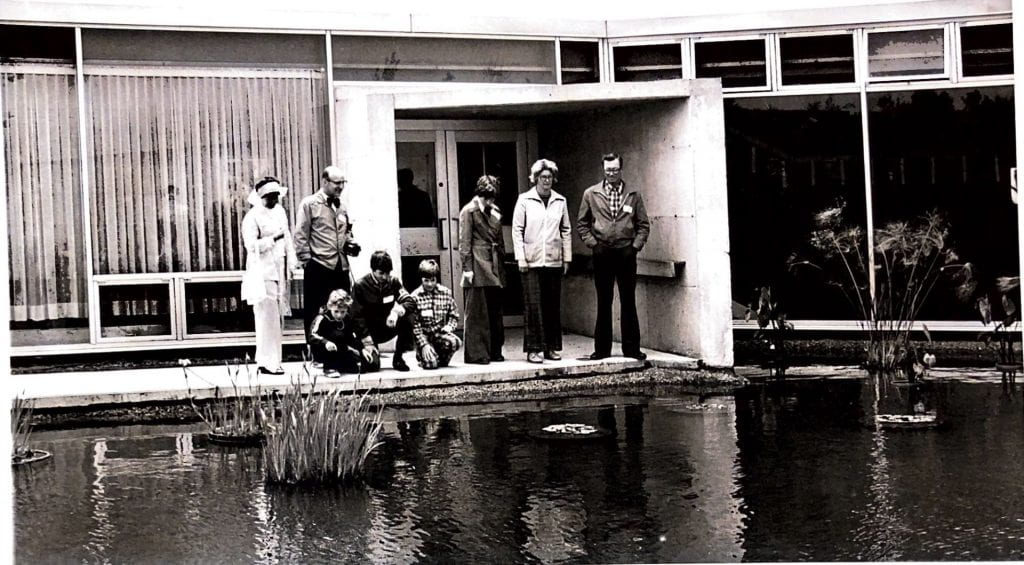 A group of adults and children stand at the edge of a pond in front of a concrete building with large glass windows. The three children kneel, looking closely at the pond. The adults stand nearby, watching the children and the pond's surface.