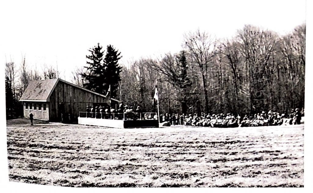 A large crowd gathers outside of a wooden building with a slanted roof. The crowd sits in chairs in rows, looking towards a stage that is bordered by flags. Behind the building and the crowd is a dense forest.