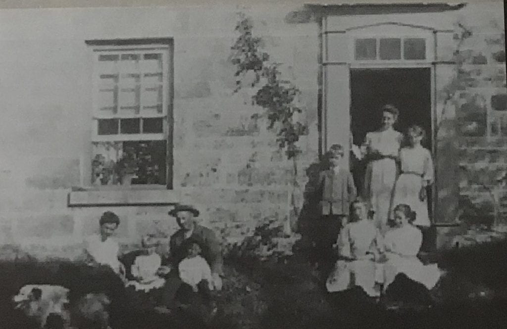 A family of nine stands in front of a brick home. Four individuals (a woman, a man, and two young children) sit on the far left underneath an open window. Five other young people stand in front of the doorway.