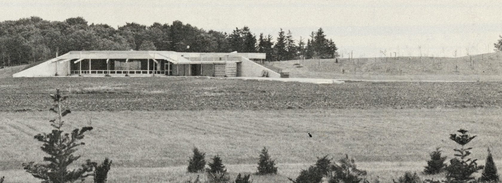A concrete building with large glass windows sits built into the side of a hill. A large open lawn rests in front of the building. Some small conifer saplings are planted into the lawn at the base of the picture.
