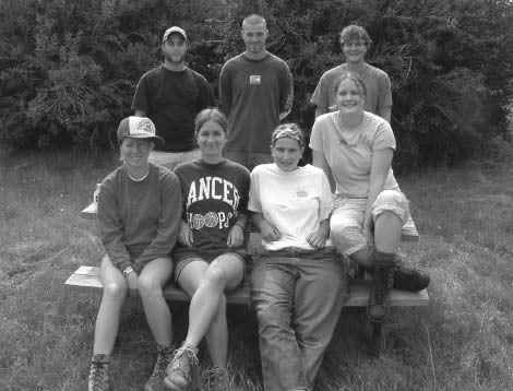 A group of seven young adults sit on a wooden picnic bench for a photo. Three men stand in the back smiling while four women sit in the front.