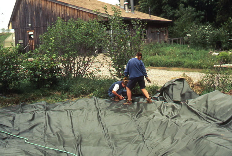 Black tarp is spread across the ground in front of a small area of bushes and a gravel road. Two individuals stand on the tarp, working. One woman is crouched over while the other person stands above them, looking down. A wooden structure with a slanted roof is seen across the gravel road behind them.