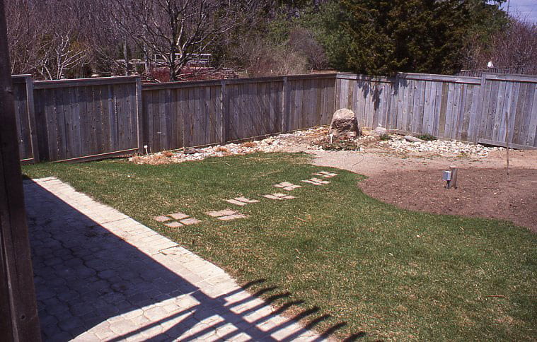 A green lawn has stepping stones to a large dirt area. A wooden fence surrounds the yard.
