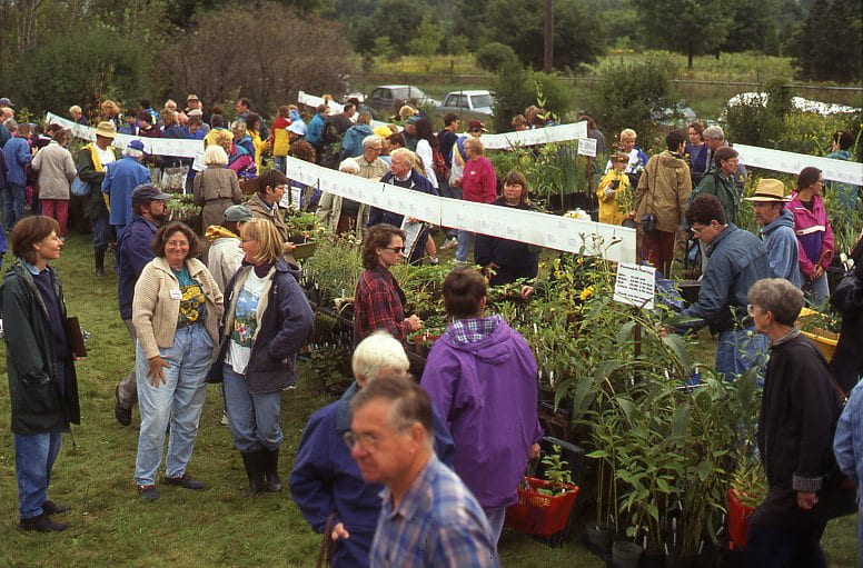 A group of people browse through rows of potted plants in a field. Informational signs are hung above the plants.