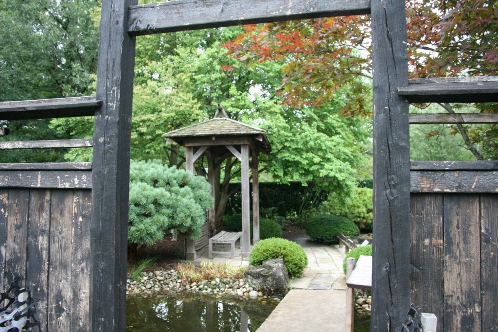 A dark wooden fence and archway surrounds a Japanese garden. Inside, a pond, wooden gazebo, bench, and many green bushes can be seen.