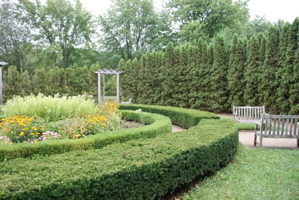 A flower bed is circled by green shrubs. To the right there are two wooden benches facing each other. Tall confiders surround the perimeter of the garden. A wooden arbor provides an entryway to the garden through the wall of conifers.
