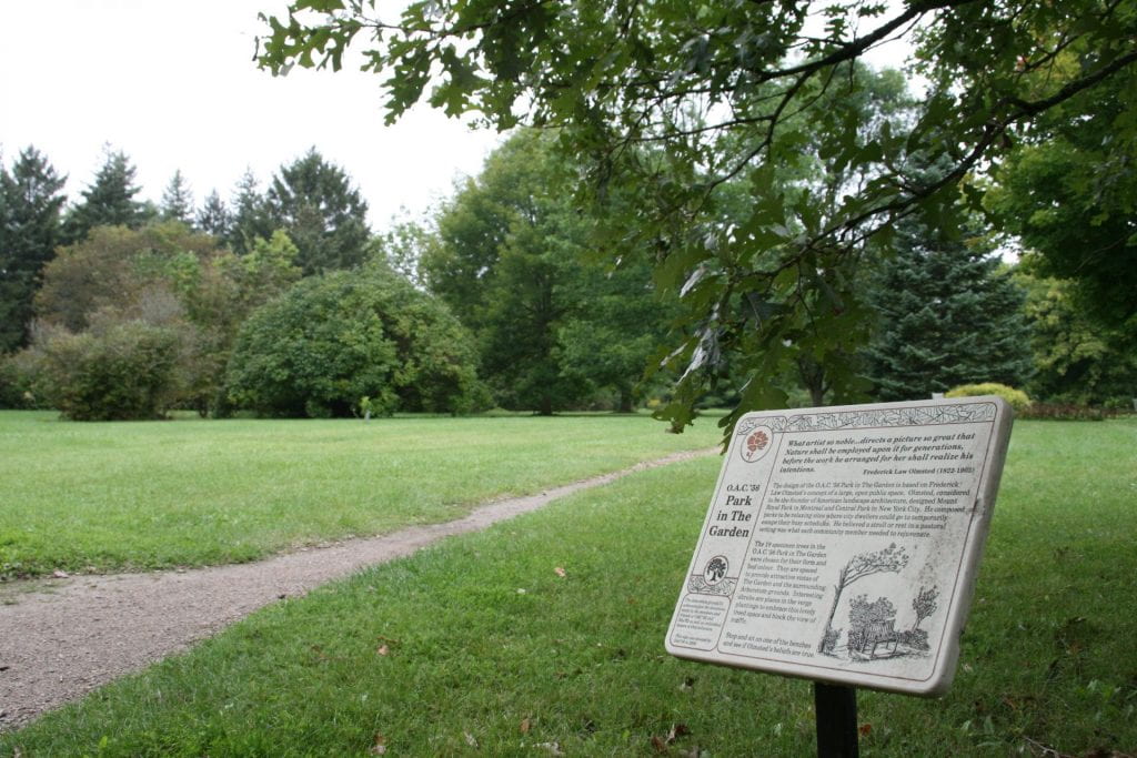 A stone plaque reading o.a.c. park in the garden is displayed bottom right of the picture, close to the ground. A dirt pathway goes left of the plaque and through the field in the distance. A densely wooded forest is seen in the background.