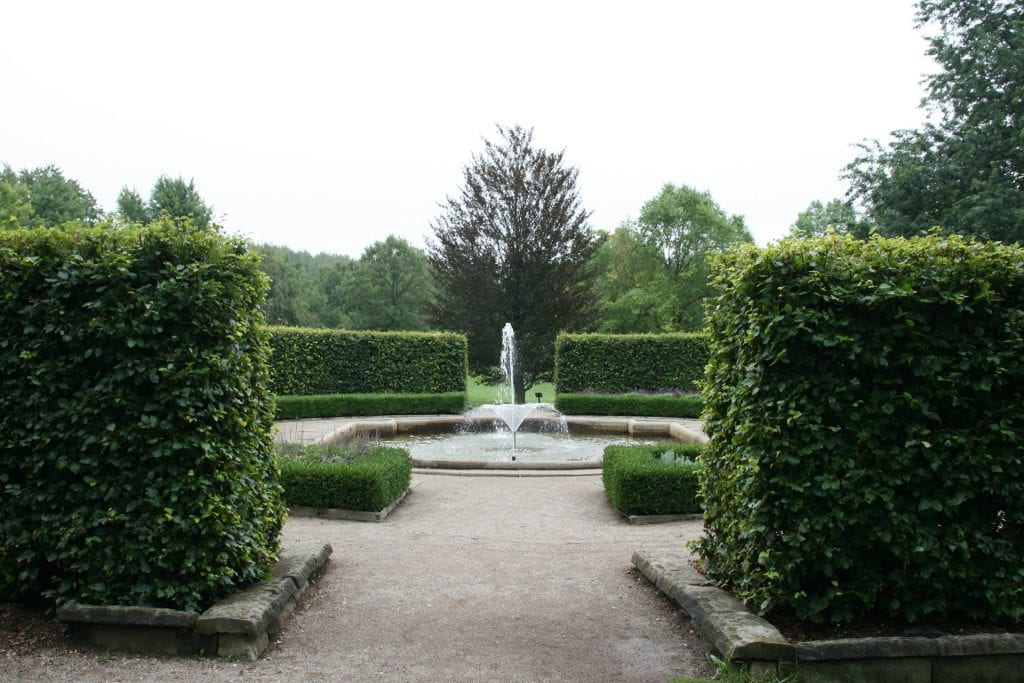 Two large green shrubs frame an entryway into a garden. A dirt path enters the garden and surrounds a pond with a large fountain in the centre. A lush green forest is seen in the background.