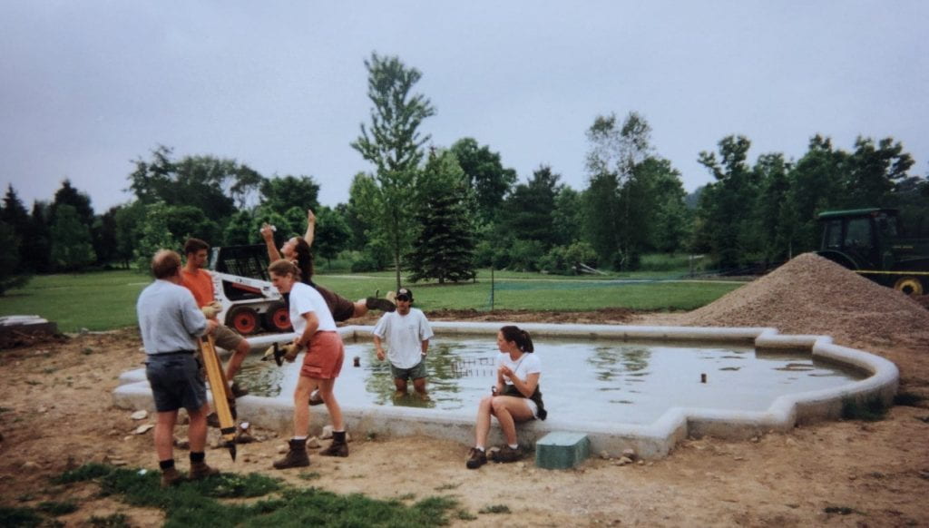 A group of staff gather around a newly placed garden pool. Most staff speak in a group around the pool. One woman sits on the edge of the pool, a man stands in the pool, and another poses with her arms outstretched on the edge. The area surrounding the pool is overturned dirt.