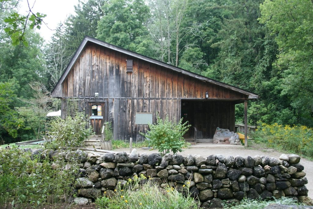 A wooden building with a slanted roof sits in front of a large forest of green trees. A wall of stacked stones sits in front of the building.