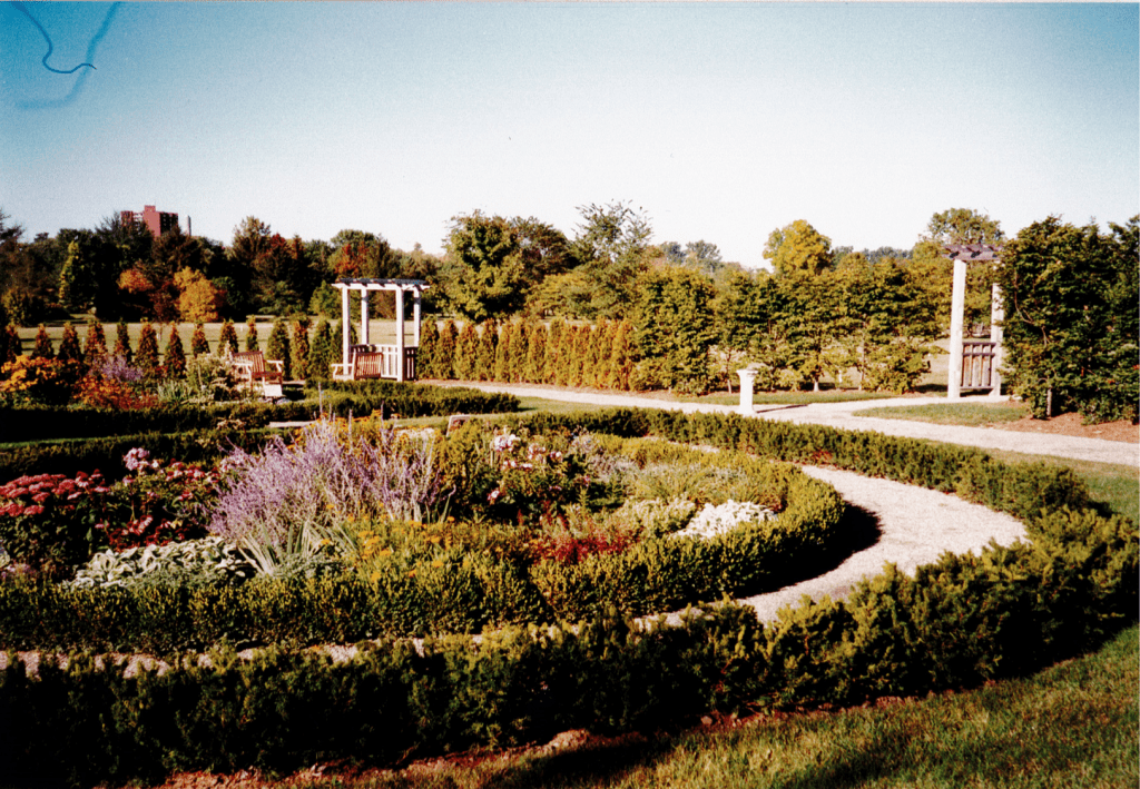 Two gardens shaped in half circles face each other. Each garden has a flower bed in the center and is outlined by rows of small green shrubs. Surrounding the gardens is a dirt path and another row of conifers. Some wooden arbors dot the row of conifers to provide entryway to the area. In the distance, a tall brown brick building can be seen above a forest.