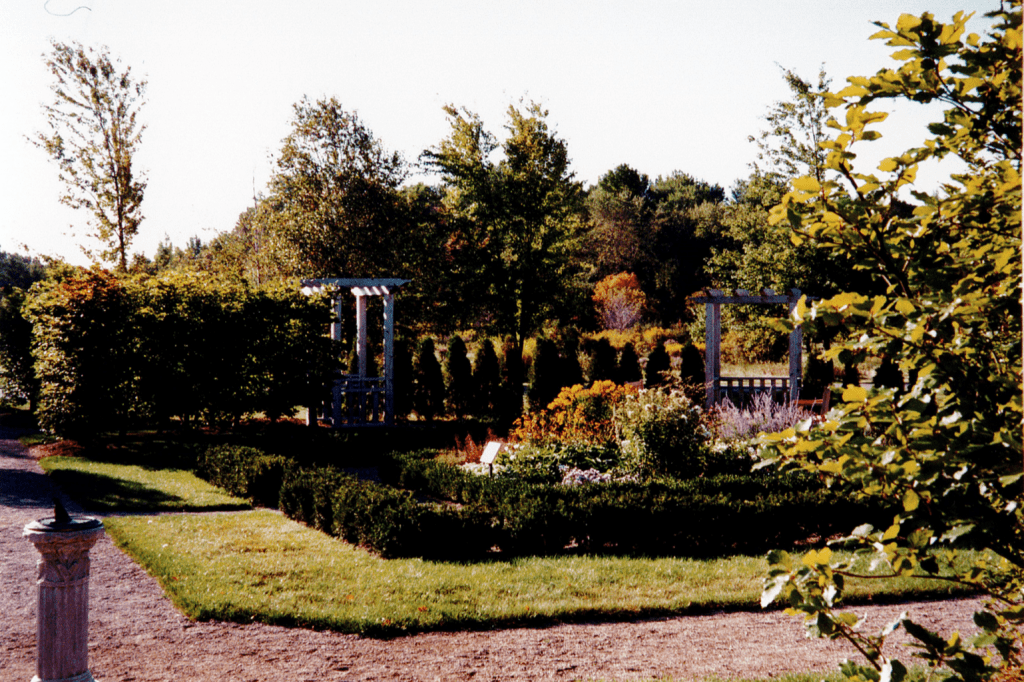 A flowerbed is circled with two rows of small green shrubs. Surrounding the shrubs is a dirt path that travels around the perimeter of the garden. On the dirt path on the left is a sundial resting on a stone column. Behind the flower bed are small conifer trees and wooden arbors providing entryways into the garden.