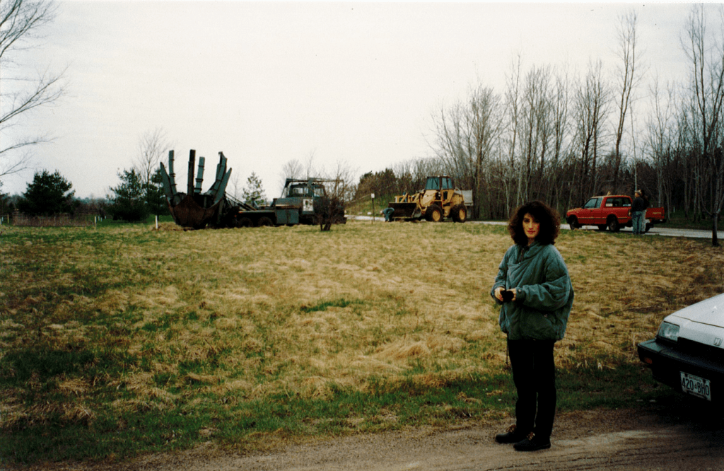 A young woman with large dark brown hair stands in front of a field with a truck and a backhoe. The woman is wearing an oversized green coat and black pants. In the distance, a truck has positioned a large tree spade in the field. A yellow backhoe and red pickup truck sits nearby.