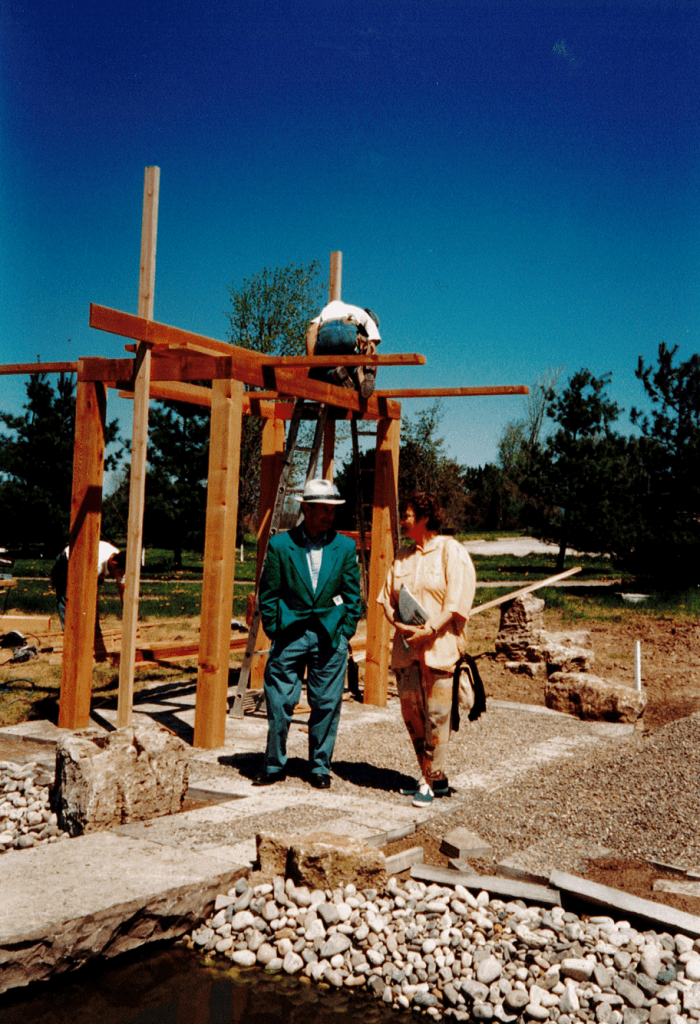 A man and a woman stand on a gravel area in front of a wooden structure being built. The man on the left is wearing a white fedora and a blue suit. The woman on his right is wearing cream button up shirt and beige pants. The two converse. Behind them, what appears to be a small gazebo is being built. A man sits on top of a ladder and is leaning over to look at where he is hammering.