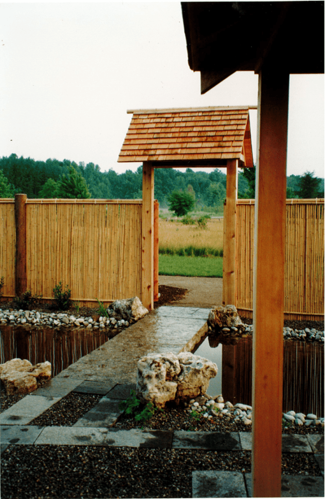A small stone pathway leads across a small pond through an archway in a bamboo fence. Through the entrance, this is an open field with a forest in the distance.