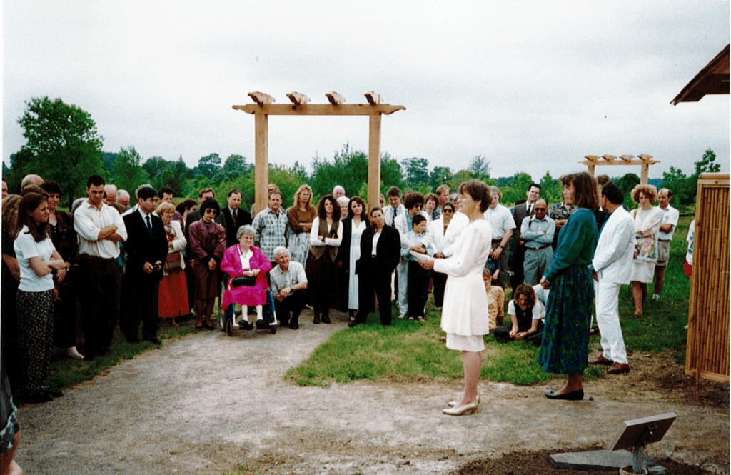 A crowd of people form a circle around two women. A woman with short brown hair is standing speaking to the crowd. She is wearing. white dress and small heels. A woman with mid-length brown hair stands behind her with her hands clasped. She is wearing a long blue dress and flats. Wooden arbors and a lush green forest can be seen being the crowd.