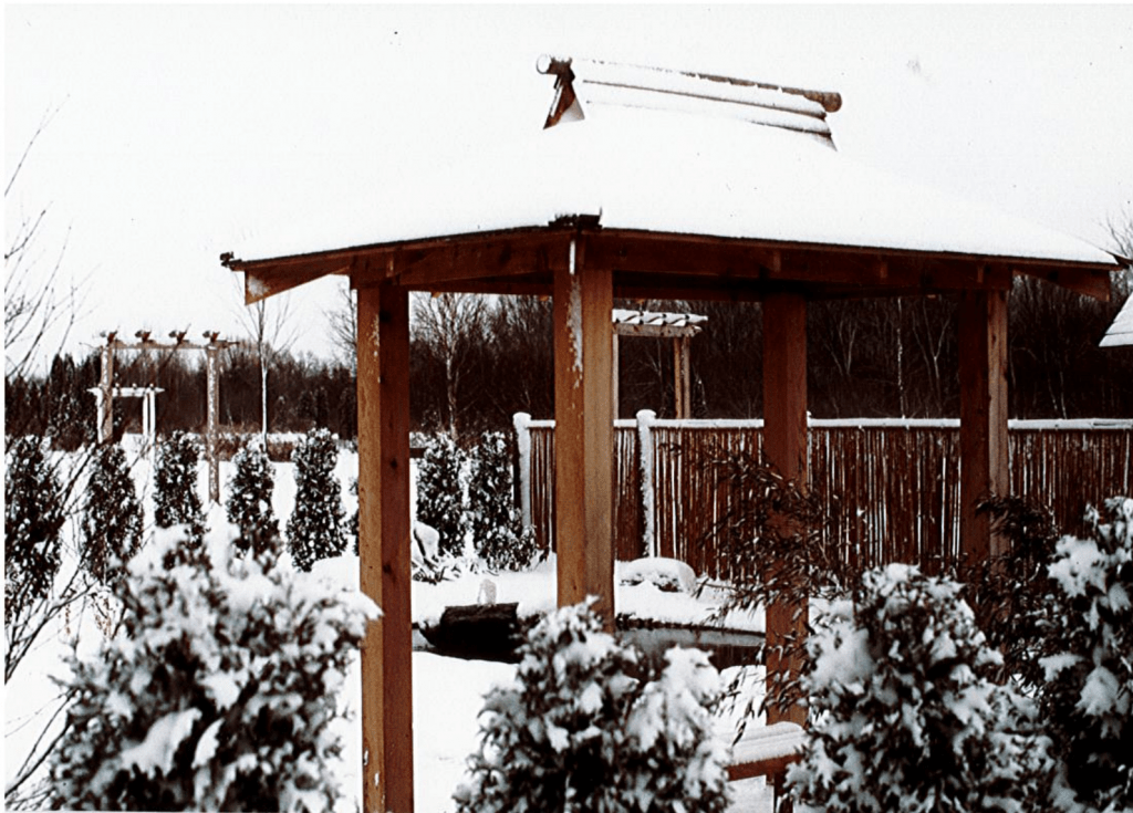 A Japanese-style gazebo stands behind a small row of snow covered shrubs. The roof of the gazebo is covered with a thick layer of fresh snow. There is a small pond, another row of shrubs, and a wooden fence behind the gazebo. Everything is covered in a fresh layer of white snow.