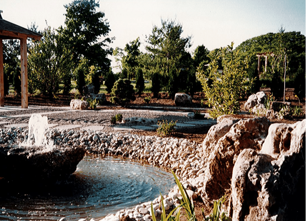 A view of a Japanese Garden. A small pond with a fountain sits in the bottom left of the frame. A groomed zen garden lies behind it. Green shrubs and small trees are planted behind the zen garden. The edge of a gazebo is seen in the far left of the picture.