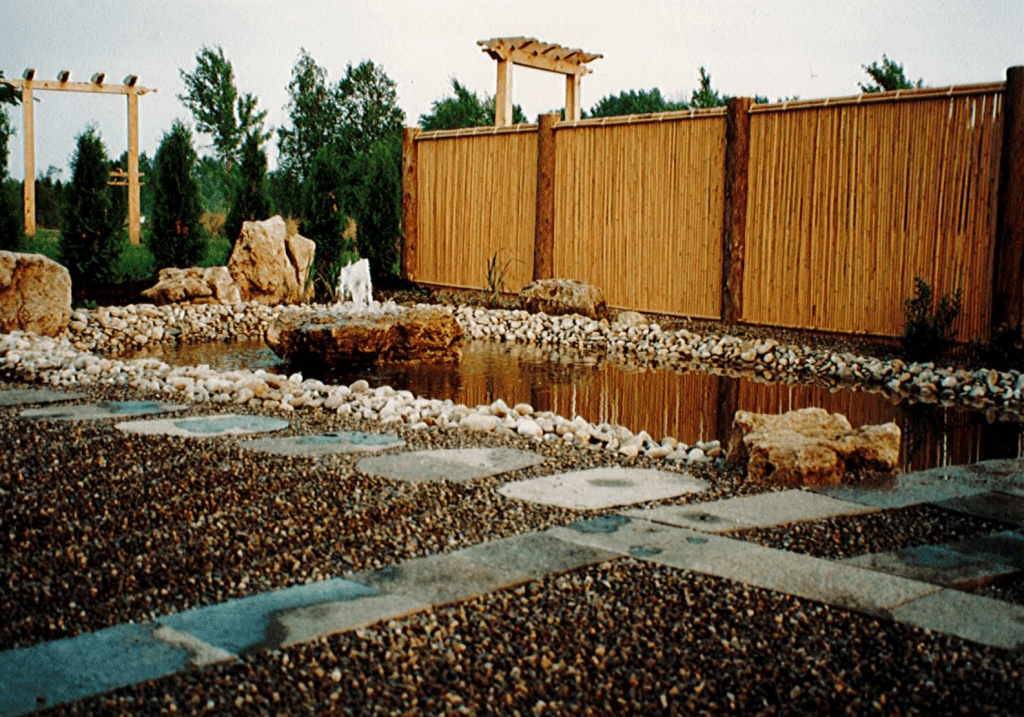 Stepping stones cross in a zen garden. A small pond with a fountain sits behind the zen garden. The pond is bordered by a tall bamboo fence. Wooden arbors and tall trees can be seen in the distance.