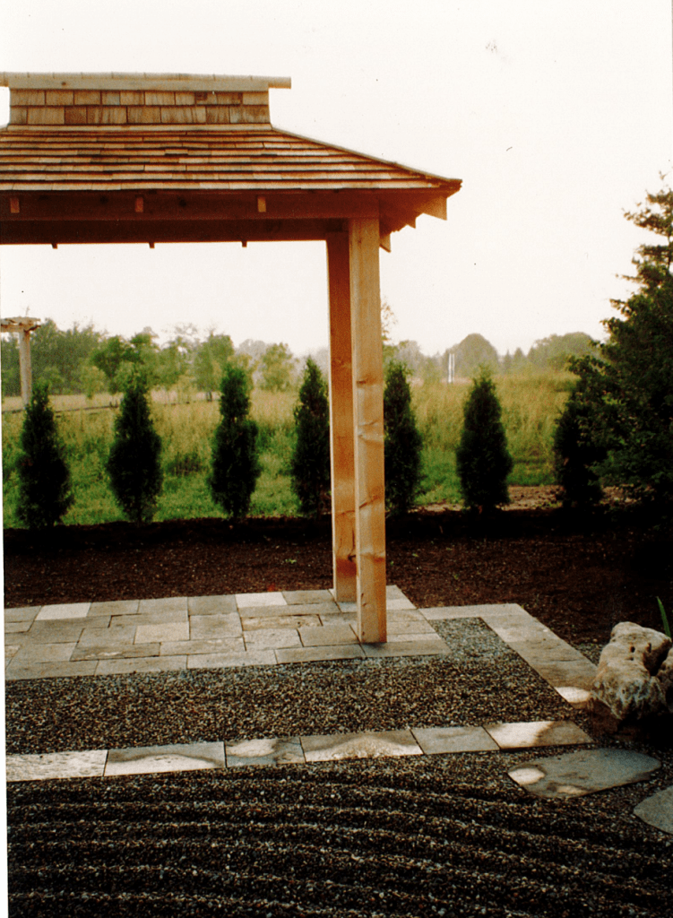A zen garden is raked to form curving lines in the grey gravel. Behind the garden sits a Japanese-style gazebo. The gazebo's wood is clearly brand new. Behind the gazebo, there is a row of small green shrubs. An open field with trees is seen in the distance.