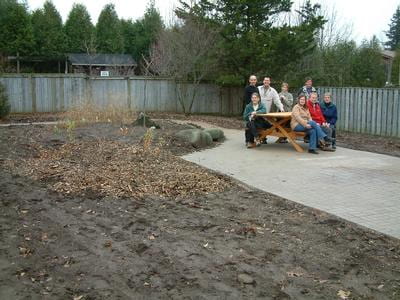 Eight people sit on a picnic table in the background. The picnic table sits on a slab of concrete in the middle of a garden. The garden surrounding them is barren, only soil that has been ploughed for future plantings.