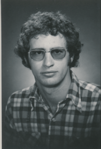 A white man with short, curly hair looks at the camera. He wears a plaid shirt and wide frame glasses.