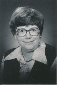 A white middle-aged woman with short hair smiles at the camera. She wears a button up shirt with a large collar, a vest, and large glasses.
