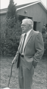 R.J. Hilton stands in a suit in front of a building. He is looking away from the camera and is holding a wooden cane in his hand.