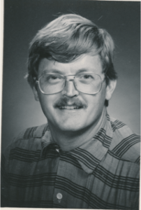 A white man smiles at the camera. He has short hair and a moustache. The man is wearing a plaid button up shirt and large wide frame glasses.