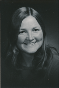 A young white woman smiles at the camera. She has shoulder length wavy hair. She is wearing a dark shirt.