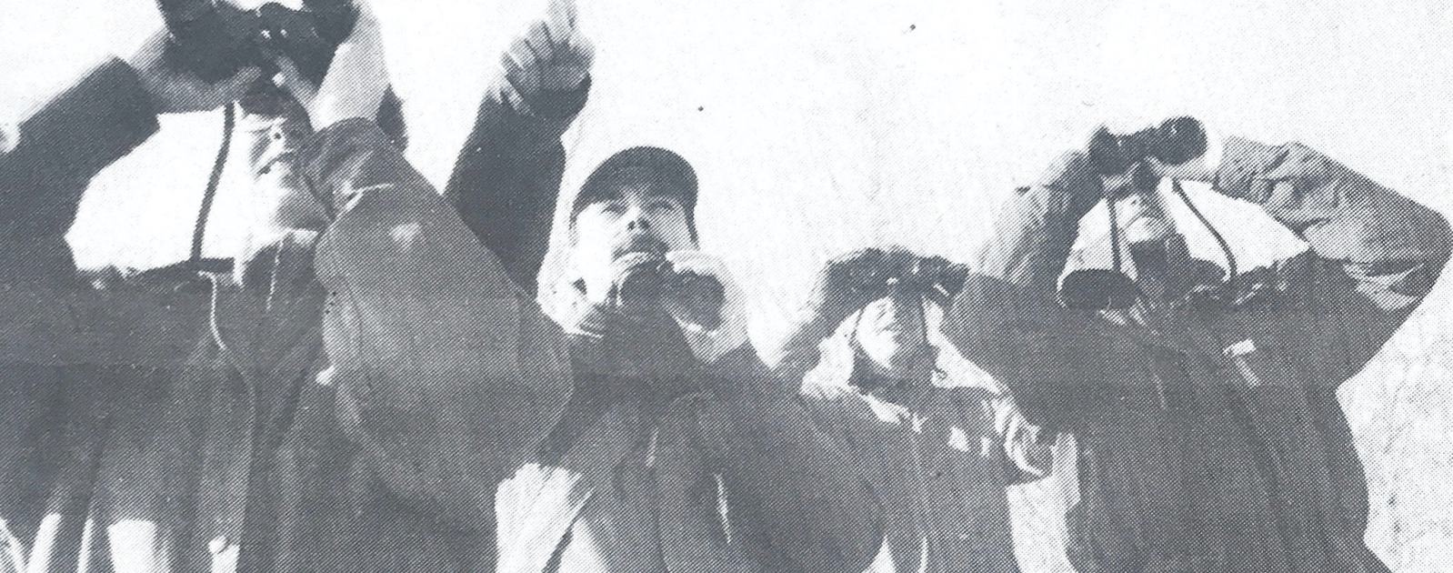 A man points in the distance while holding binoculars. The three individuals surrounding him look through binoculars in the direction he is pointing.