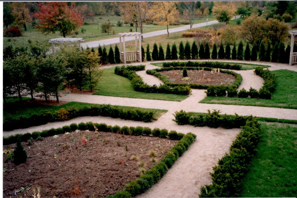 A garden surrounded by green shrubs and trees. In breaks in the trees are wooden arbors leading in to the garden. Paths weave through the garden. The flower beds are occupied by small sprouts of plants.