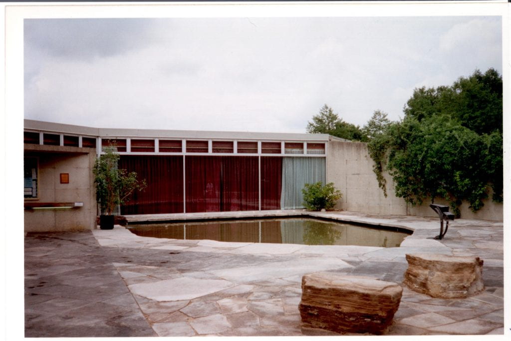 A concrete building with large windows has a small pond in front of it. The pond is surrounded by concrete. Two stones and a metal statue of a bird are placed near the pond.