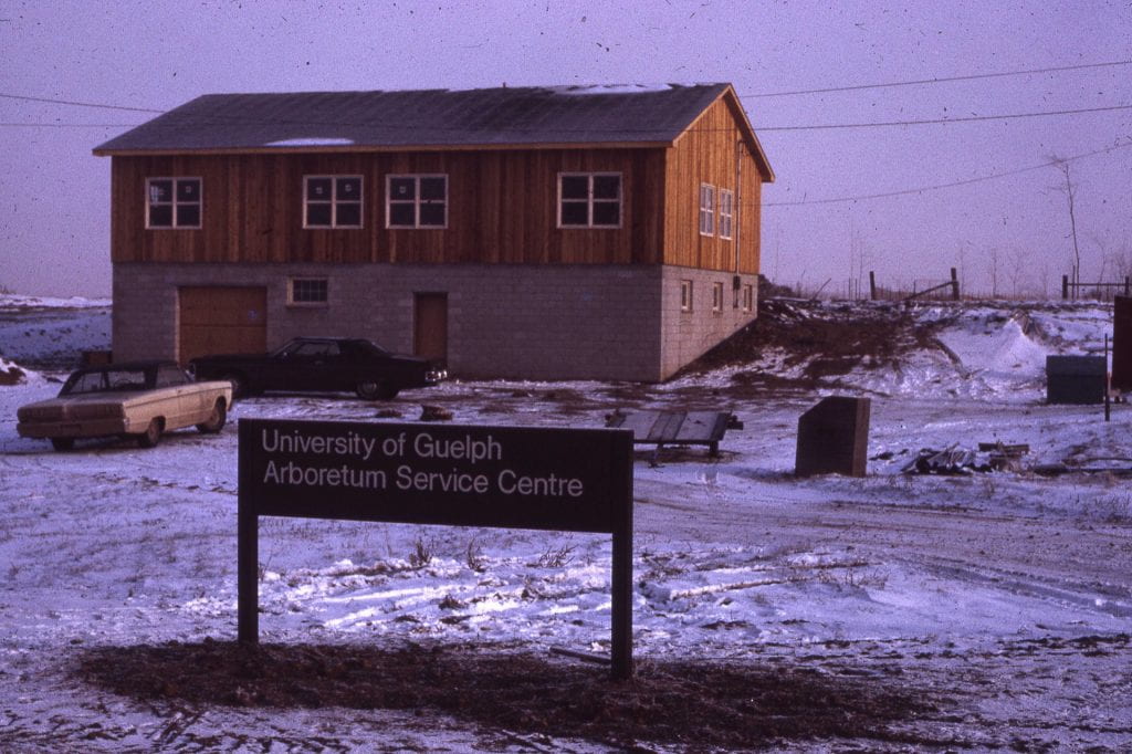 In the background, a wood and brick two-level building stands surrounded by snow. Two cars are parked in front. A sign reads "University of Guelph Arboretum Service Centre" in front.