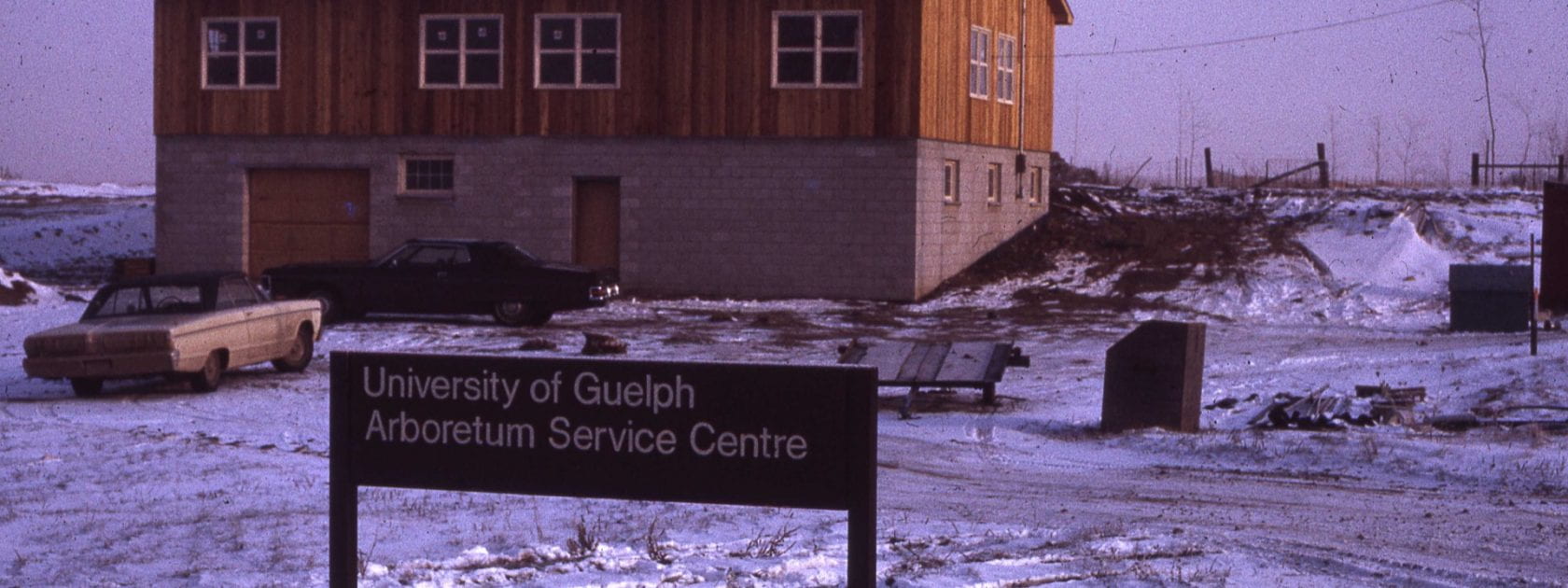 In the background, a wood and brick two-level building stands surrounded by snow. Two cars are parked in front. A sign reads "University of Guelph Arboretum Service Centre" in front.