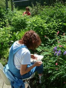 A woman with brown, curly hair sits on a chair facing towards a flower bush with pink and purple blooms. She looks closely at the objects in her hand while she prepares to paint them with a watercolour kit resting on her lap.