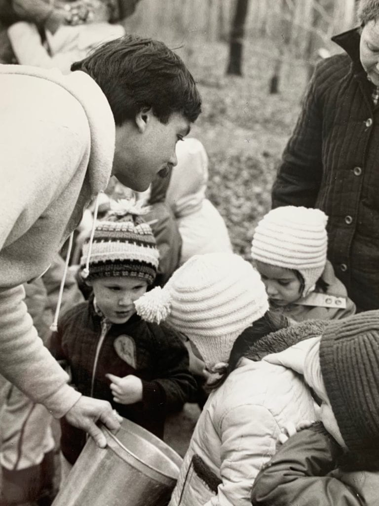 A man with short dark hair leans over, showing the contents of a silver bucket to a group of young children.