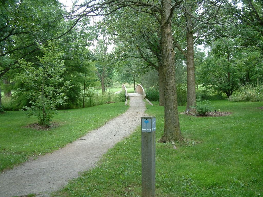 A dirt trail leads towards a small bridge. There is green grass and large trees with green leaves on either side of the trail.
