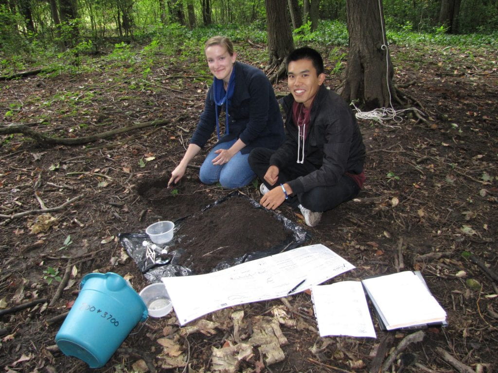 Two students, a young woman and a young man, kneel on the forest floor to dig for invertebrates. They are smiling at the camera.