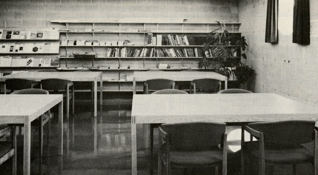 Four tables with chairs are spaced around a room. On the back wall, a wall-to-wall bookshelf is filled with books. A large plant sits in the back right corner of the room.